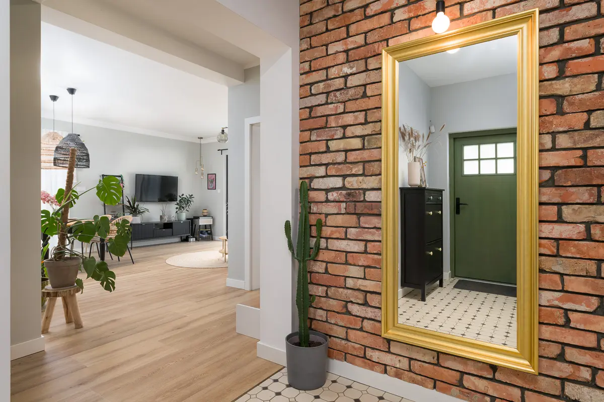 Stylowy przedpokój z lustrem w złotej ramie i ścianą z naturalnej cegły.